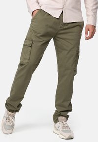 Pantalones cargo verde olivo de algodón, con bolsillos laterales, corte recto y dobladillos ajustados, combinados con zapatillas de color claro.