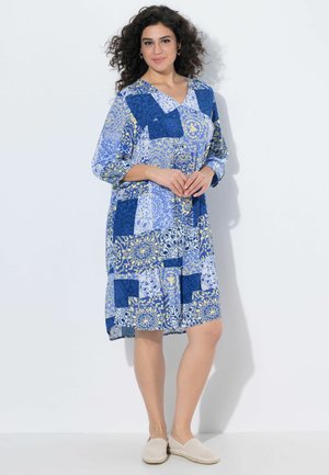 Kvinde står smilende iført en knælængde blå og hvid patchwork mønstret kjole med 3/4 ærmer og beige slip-on sko mod en hvid baggrund.