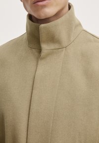 Cappotto beige con colletto alto, realizzato in tessuto texture. Presenta un design snello con cuciture minime e una finitura morbida.