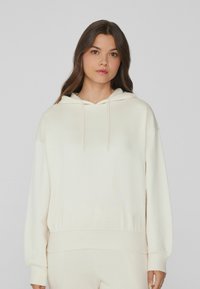 Helle Kapuzenjacke aus weichem Stoff. Verfügt über eine Kordelzugkapuze, gerippte Bündchen und einen lockeren Schnitt. Nahtloses Design mit minimalen Details.
