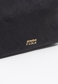 Bolsa de tela negra con una textura suave; presenta un logo dorado en relieve "BIMBA Y LOLA" en el frente. Diseño y forma minimalistas.