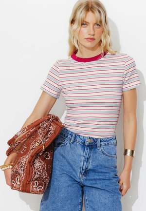 Femme aux longs cheveux blonds porte un haut blanc à manches courtes avec des rayures rouges, un jean bleu taille haute, et porte un sac marron à motif sur un bras.