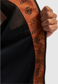 Sort mesh-stof med en orange satinforing, prydet med et trykt mønster af stjerner og tal, samt initialerne "FB" i sort.