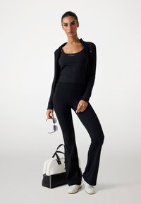 Black athletic outfit featuring a long-sleeve zip jacket, fitted tank, and flared pants. Includes white and black handbag and sunglasses.