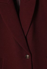 Dvojvrstni plašč v burgundski volneni mešanici. Ima zareznje obroče in en sam gumb kot poudarek. Tekstura tkanine z gladkim zaključkom.