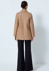 Beige læderjakke med lige design, notch krave og lange ærmer, parret med vide sorte bukser. Bagudsigten er synlig.