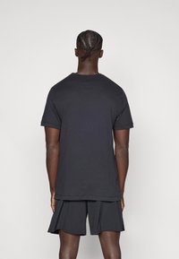 Μαύρο βαμβακερό μπλουζάκι με κοντά μανίκια, άνετη γραμμή και στρογγυλή λαιμόκοψη, συνδυασμένο με μαύρα σορτς σετ, εμφανιζόμενο από πίσω.