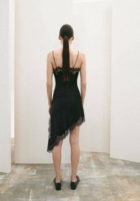 Vestido negro asimétrico con ribete de encaje, tirantes finos y textura sedosa. Combinado con zapatos negros sin cordones, visto desde atrás.