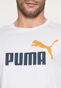 Balta medvilninė marškinėliai su dideliu tamsiai mėlynu "PUMA" logotipu ir oranžiniu šokinėjančio pumos grafiku virš teksto. Klasikinis apvalios kaklo dizainas.