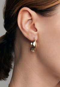 Złote kolczyki w kształcie obręczy o gładkiej, wypolerowanej powierzchni, charakteryzujące się wydłużonym, asymetrycznym kształtem i małym detalem akcentującym.