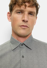 Mann mit heller Haut und lockigem braunem Haar, der ein graues kariertes Hemd mit Knöpfen trägt und mit neutralem Gesichtsausdruck geradeaus blickt.