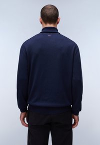 Middengrijze pullover trui met een hoge kraag, geribbelde manchetten en zoom. Heeft een minimalistisch logo detail op de rug nabij de nek.