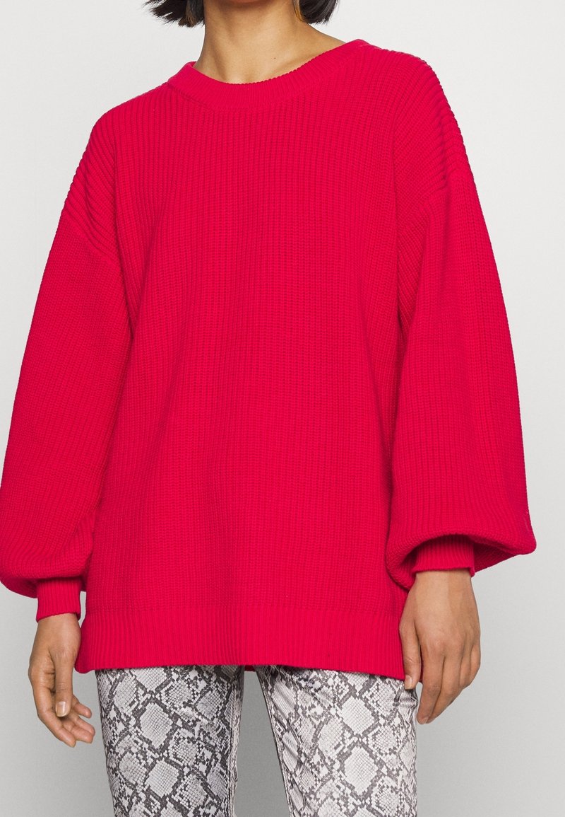 Femme portant un pull en tricot rouge vif, ample, avec des manches froncées et un pantalon à imprimé serpent taille haute sur un fond uni.