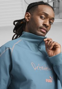 Gaiši zils, augsta apkakle, džemperis ar rozā uzrakstu "Noteikti" un Puma logo; ietver rāvējslēdzējus, ribotas manšetes un šuvju detaļas.