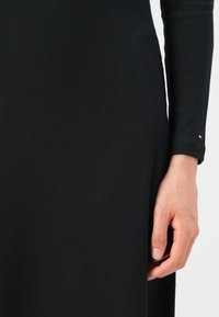 Robe noire côtelée avec manches longues, une silhouette ajustée et un petit logo près du poignet. Texture lisse sur l'ensemble.