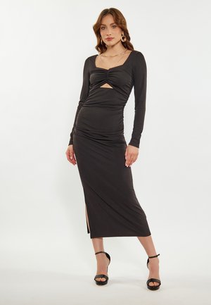 Frau trägt ein langes schwarzes figurbetontes Kleid mit langen Ärmeln, gerafftem Oberteil, Taillenausschnitt, seitlichem Schlitz und schwarzen Sandalen mit hohem Absatz.