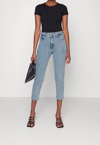 Mujer con pantalones vaqueros cortos de tiro alto en un azul claro, camiseta negra ajustada de manga corta, tacones destalonados negros, sosteniendo un bolso de mano negro.
