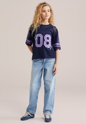 Fiatal személy hosszú, göndör hajjal, sötétkék, túlméretezett "08" számot és csillagokat ábrázoló ingben, világoskék farmerral és kék tornacipővel, lazán állva.