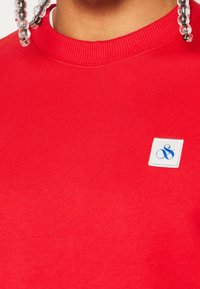 Sudadera roja con cuello redondo acanalado. Presenta una pequeña etiqueta blanca de tela con un logo azul. Textura suave, diseño casual.