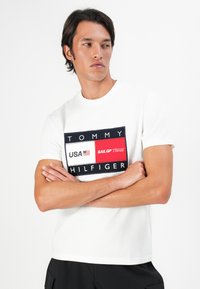 Homme debout, bras croisés, portant un t-shirt blanc de l'équipe SailGP de Tommy Hilfiger et un pantalon noir, sur fond blanc.