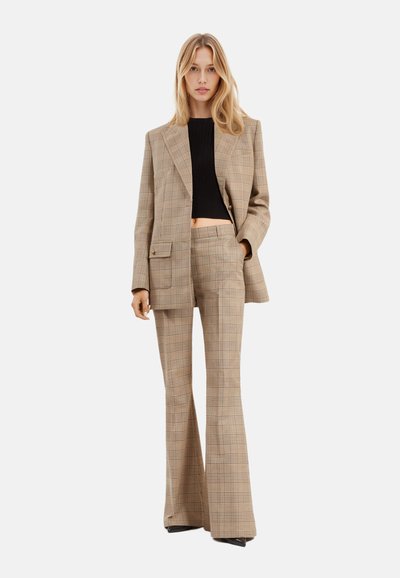 Costume écossais beige avec une veste croisée et un pantalon évasé. La veste est ornée d'une poche et de détails dorés. Top noir court en dessous.