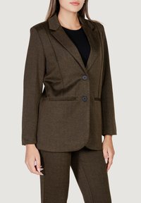 Blazer de espiga marrón con dos botones frontales, solapas con muesca y bolsillos laterales, combinado con pantalones a juego texturizados.