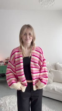 Νεαρή γυναίκα μέσα σε εσωτερικό χώρο, φοράει πουλόβερ με ροζ και λευκές ρίγες πάνω από μαύρη μπλούζα και μαύρο παντελόνι, στέκεται σε ένα άνετο σαλόνι.