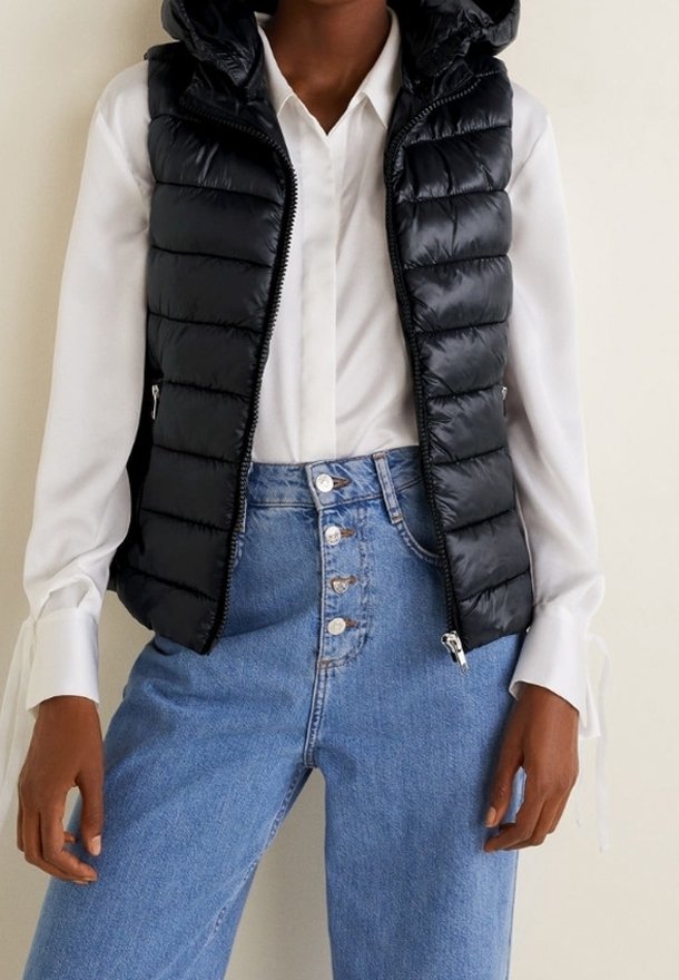 Černá prošívaná péřová vesta s kapucí, s horizontálním prošíváním, zapínáním na zip a bočními kapsami, nošená přes bílou košili s límečkem a džíny s vysokým pasem.