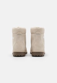 Botas de tobillo de ante beige con una parte superior redondeada, detalles de costura y una suela de goma marrón. Presenta un logo en el lado exterior. Acabado suave.