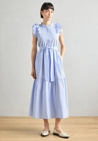 Gestreiftes Midikleid in Blau und Weiß mit Rüschensäumen, Knopfleiste, tailliertem Bund, geschichtetem Rock und Taschen. Trägt flache Schuhe.