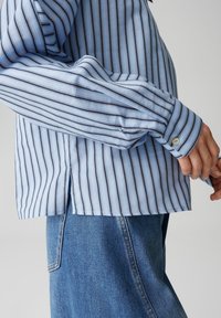 Chemise à rayures bleu clair avec des lignes verticales en bleu foncé et blanc. Manches longues avec poignets boutonnés et fentes latérales à l'ourlet.