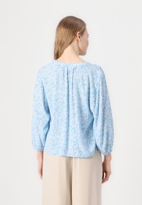 Blusa azzurro chiaro con motivo floreale bianco, scollatura rotonda e dettagli arricciati sul retro. Maniche lunghe con polsini elasticizzati.