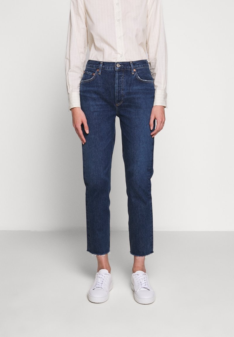 Personne portant un jean droit bleu foncé avec des ourlets effilochés, une chemise blanche à manches longues boutonnée et des baskets blanches.