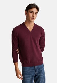 Giovane uomo che indossa un maglione bordeaux a V e jeans blu, in piedi con una mano dietro la schiena su uno sfondo bianco.