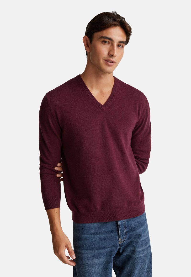 Giovane uomo che indossa un maglione bordeaux a V e jeans blu, in piedi con una mano dietro la schiena su uno sfondo bianco.
