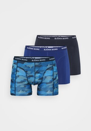 Trīs vīriešu bokseršortus pāri: viens tumši zils, viens viendabīgs zils un viens zils kamuflāžas raksts. Visām ir melna josta ar logotipu.