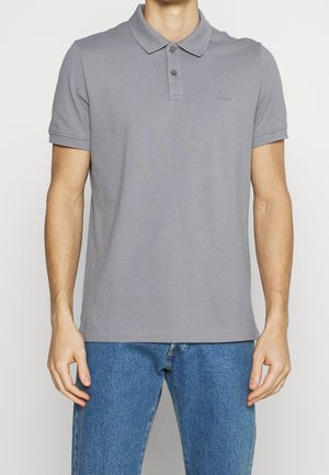 Mężczyzna ubrany w prostą szarą koszulkę polo z krótkim rękawem oraz niebieskie dżinsy, stojący na tle jednolitego jasnego tła.