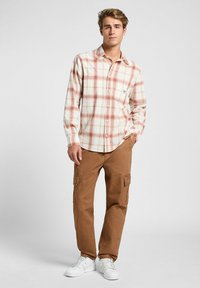 Camisa de cuadros en beige con acentos en rojo y azul, combinada con pantalones cargo marrones y zapatillas blancas. Presenta un bolsillo en el pecho y mangas largas.