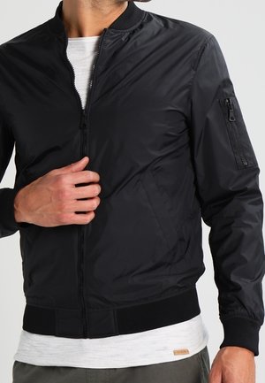 Chaqueta bomber negra con cierre de cremallera, puños acanalados y un bolsillo con cremallera en la manga izquierda. Textura suave y material ligero.