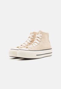 Beige High-Top-Sneaker aus strukturiertem Canvas, mit cremefarbenen Schnürsenkeln, einer weißen Gummisohle und einem schwarzen Streifen an der Zwischensohle.
