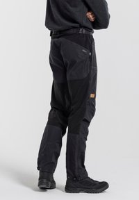 Black outdoor pants with a relaxed fit, made of durable fabric, featuring zippered pockets and reinforced knees. Paired with black footwear.