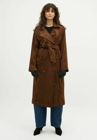 Bruine suedetrenchcoat met een ceintuur in de taille, dubbele knopen en grote knopen; gedragen over donkere jeans en zwarte hakken.