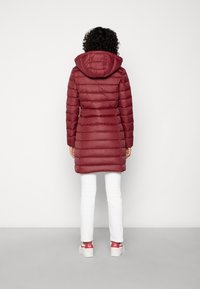 Abrigo acolchado burdeos con capucha, textura acolchada y costuras verticales. Se lleva con pantalones blancos y zapatillas con detalles en rojo.