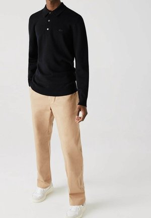Mężczyzna ubrany w czarną, długorękawową koszulkę polo, beżowe spodnie i białe trampki, stojący na tle białego tła.