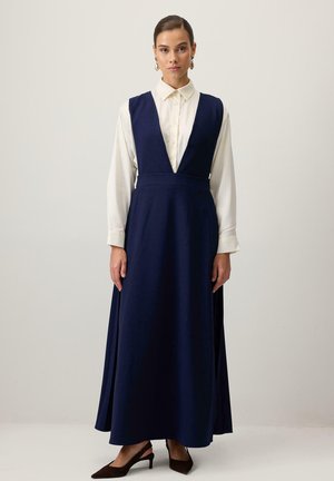 Rochie bleu marin, fără mâneci, cu decolteu adânc în V, talie strânsă și fustă fluidă. Pur și simplu pe un cămașă albă cu guler și asortată cu pantofi cu toc.