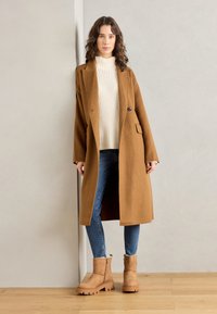 Abrigo de lana marrón con un solo botón y bolsillos laterales, combinado con un jersey de cuello alto crema, jeans azules y botas de tobillo beige con suela gruesa.