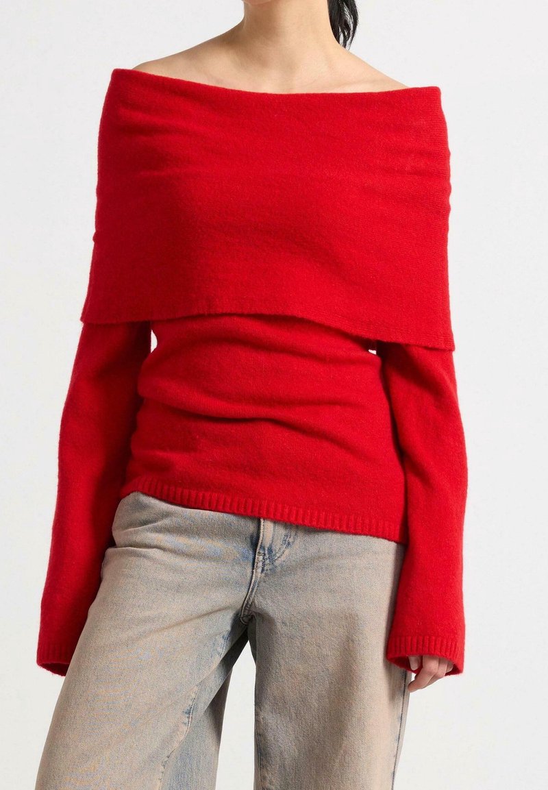 Rød off-the-shoulder sweater lavet af blødt stof med lange ærmer og et lagdelt design. Kombinationen med lyseblå jeans.