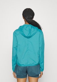Giacca a vento color teal con cappuccio, texture liscia e polsini elasticizzati, abbinata a pantaloni corti verde scuro, vista da dietro.