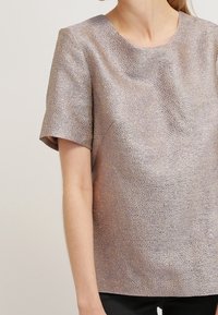 Žena v krátkém rukávu s texturovaným metalickým béžovým topem s kulatým výstřihem, doplněná černými kalhotami na světlém jednobarevném pozadí.