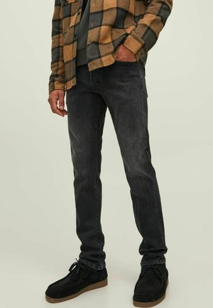 Vaqueros de denim negro con un corte ajustado, combinados con una camisa de franela a cuadros en tonos marrón y negro. Zapatos de ante negros y un suéter gris debajo.